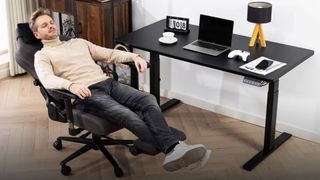 Masc presenting person napping on gaming chair next to Boulies MagVida desk with laptop and controller next to tea cup, lamp, and flip calendar 